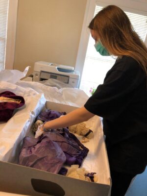 A young woman wearing gloves touching a purple antique dress in a large box surrounded by tissue paper