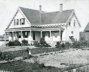 A two-story house with front porch in a yard with a garden and two figures standing in the grass
