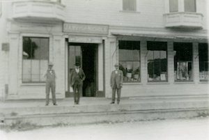 Three men standing outside of a large building with sign above the door "Jarvis & Nichols"