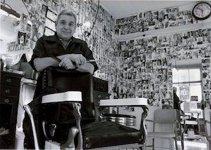 An older man standing behind a barber's chair in a shop with walls covered in photographs