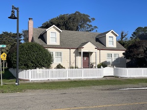 Small house with trees on either side and a white fence in front