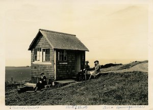 A small wooden building on a grassy hillside with three people outside it
