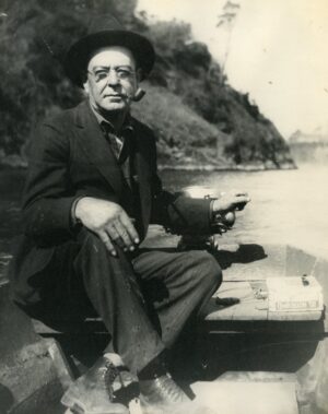A bespectacled man in a suit and a hat smoking a pipe while steering a small boat on the water with trees on the shoreline behind him