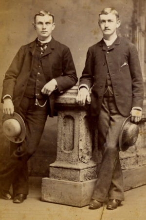 Studio portrait of two men in suits holding their hats leaning against a plinth