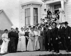 A large group of well-dressed men and women posing in front of a Victorian house
