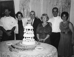 An elderly couple standing between two other couples behind a table featuring a tall decorated cake