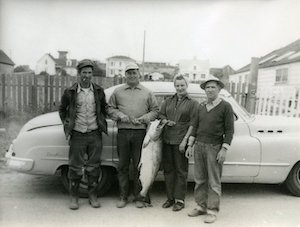 Three men and a woman posing with a large fish in front of an old car
