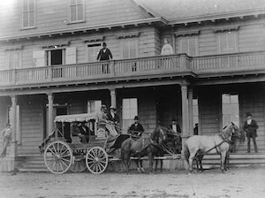 A stagecoach pulled by four horses with two drivers in front of a large building with a second floor balcony