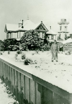 Man in a snow-covered yard with a large Victorian house, water tower, and trees in the background