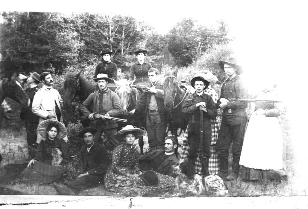 A group of men, women and a child with horses and dogs pose in a field with trees in the background. Two of the men hold rifles.