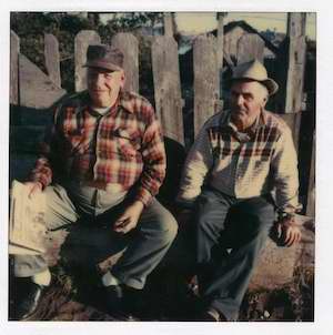 Two older men sitting in front of a wooden fence
