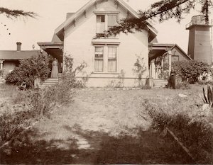 Two story home with porches on either side and a woman standing on one of them. A water tower is in the back