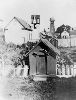 A small wooden shed with fence, yard, buildings and water tower behind it
