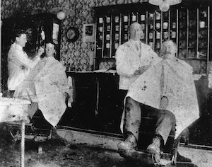 Two seated men getting hair cut by barbers in a shop