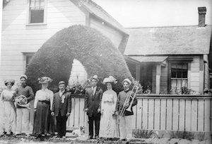 Well-dressed people, some holding musical instruments, standing in front of a fenced-in front yard and house