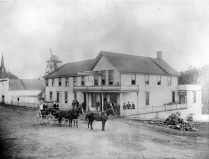 Horse-drawn wagon on a dirt street in front of a large two-story building