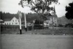 Intersection of Main and Lansing Streets, 1957. Traffic signs at the intersection of Main and Lansing Streets in Mendocino, looking eastward. At the time this photo was taken, Lansing Street was Highway 1 through town. The pine tree visible on the right side in this intersection was planted by Dr. Russell Preston (1878-1954), and in 1965 it became the community Christmas tree with lights and ornaments. The triangle around it would be landscaped the next year by the Presbyterian Youth Fellowship in memory of Mike Hill, who was lost to the ocean while surfing on January 17, 1966. The board fence encloses empty lots owned at this time by the Kelley Estate. They would later become Rotary Park in 2013. The white house in the background on the left is the Lansing House. To the right is Schlafer's Garage, built in 1914, with its corrugated metal roof. The steeple of the Mendocino Presbyterian Church, built in 1867, is visible on the right. (Gift of Bette Duke, Robert R. Duke Collection, Kelley House Photographs)