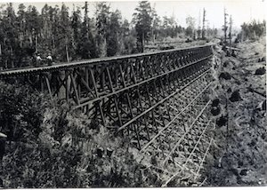 Two women and a man walking across a wooden railroad trestle above a creek