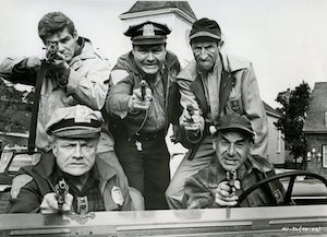 Five male actors in a car, facing camera with guns drawn