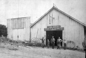Men and boys stand in front of a blacksmith shop with wagon wheels in the yard