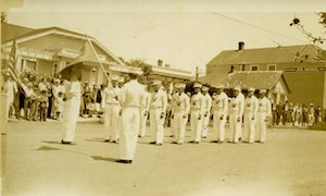 Male drill team standing on a street lined with buildings and spectators