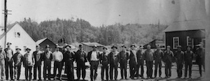 A line of men standing outside at a lumber mill, with trees and buildings in background