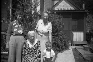 Two women and a toddler stand around an elderly woman outside a house