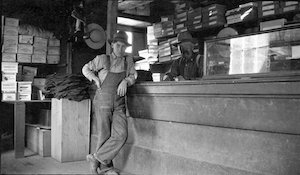 Henry Reed Ferrill and son, John, in his general merchandise store in Albion, 1910 - 1915. A pile of pants are folded behind John and father, Henry, stands behind the counter. A wide brim hat hangs on the wall and several boots hang from the ceiling.  Numerous shoe boxes are stacked in the background, some made by "Buckingham & Hecht,” a large San Francisco shoe manufacturer. (Gift of Jim and Judy Tarbell, Lloyd Henry Ferrill Collection, Kelley House Photographs)