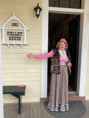 Woman in a long skirt standing on a porch in front of a sign that reads "Kelley House"