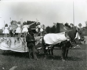 Little girls dressed as angels on a parade float, with man holding the reins of the horses.