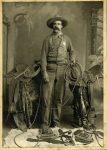 Man dressed as a cowboy/sheriff with a background of horse saddles, ropes and spurs.