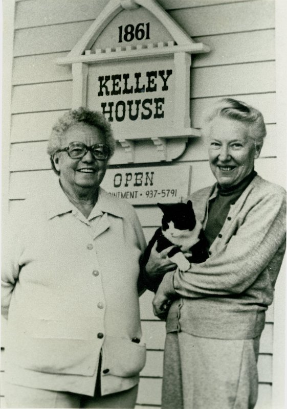 Two women stand in front of a sign “1861, Kelley House.” One holds a black and white cat.