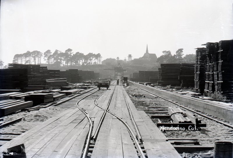 Train tracks running through lumber yard with stacks of lumber on both sides of tracks. A rail car and crane can be seen in background