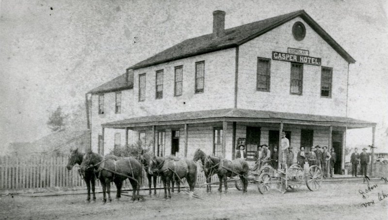 Two-story hotel building with men standing outside. A wagon pulled by six horses is in the street in front of the hotel.